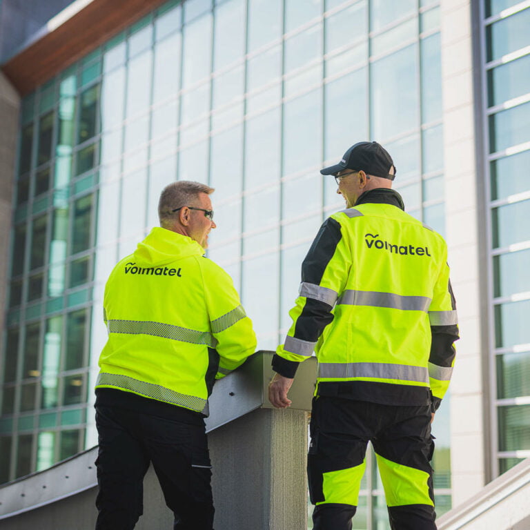 Kaksi miestä Voimatelin työvaatteissa seisoo ja keskustelee ulkona.